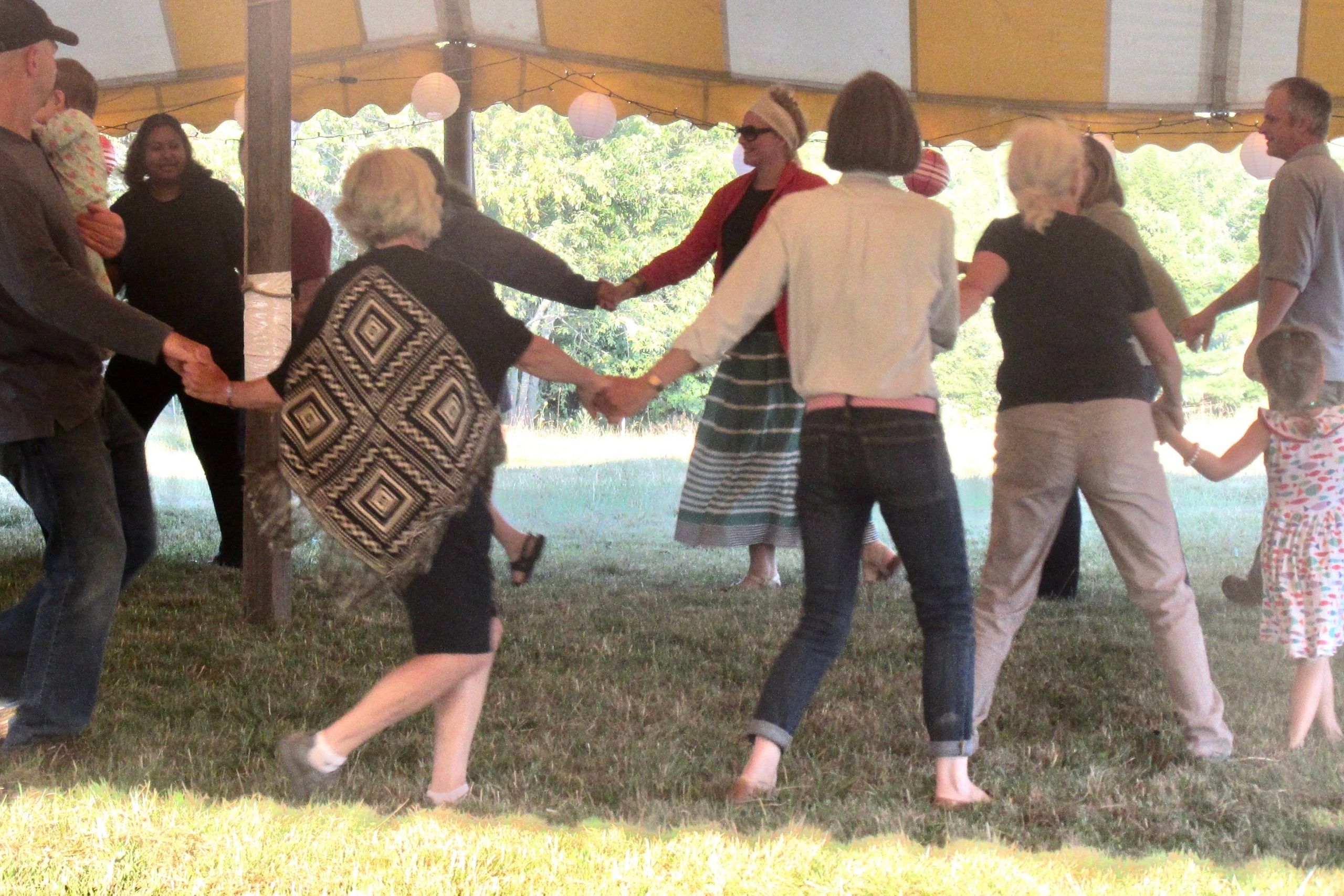 people of all ages at a farm event in Midcoast Maine dancing holding hands and dancing in a circle under a yellow and white striped tent.