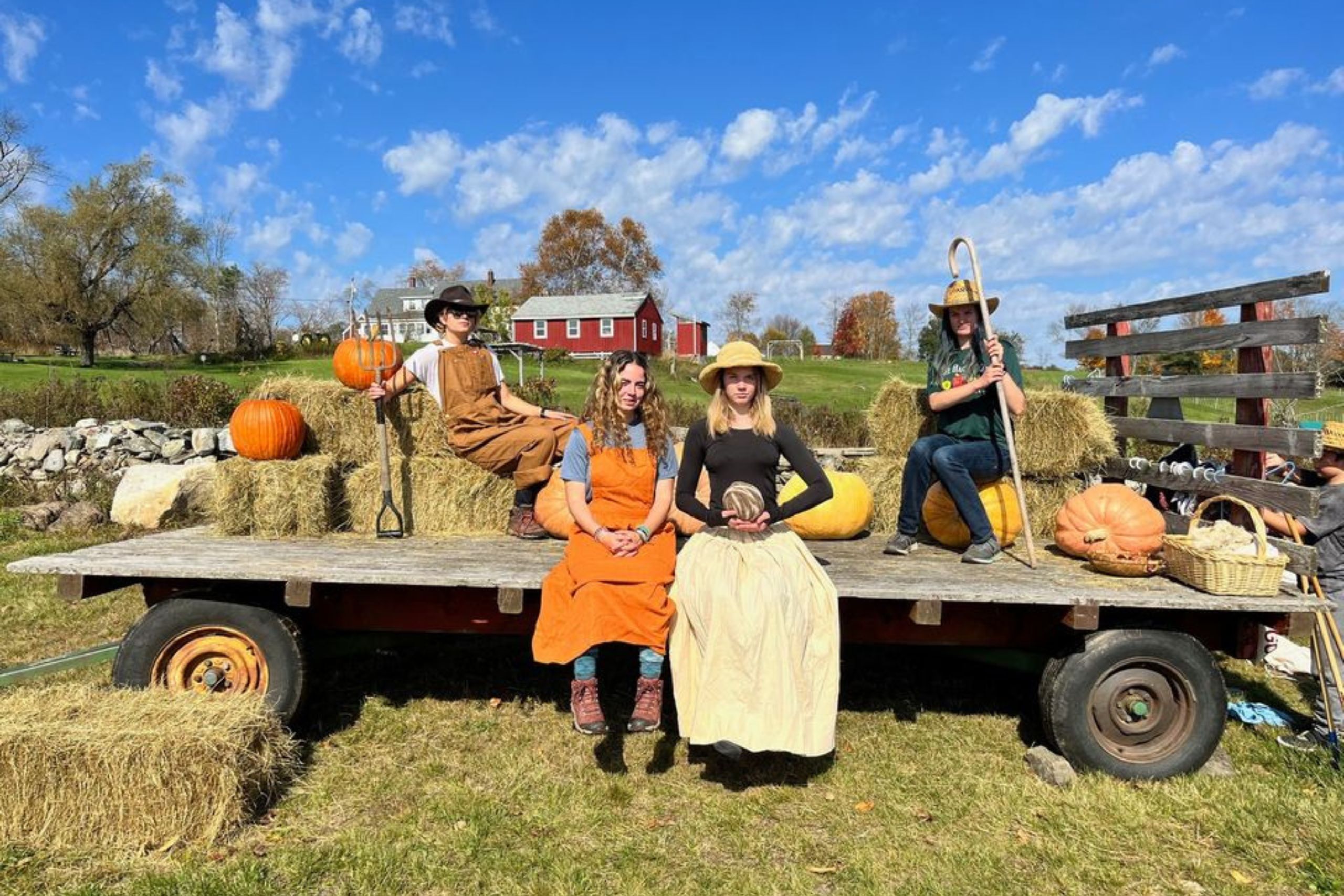 Four people in farmer dress up costumes on a hay wagon with pumpkins under a blue sky with clouds and a red barn behind them.