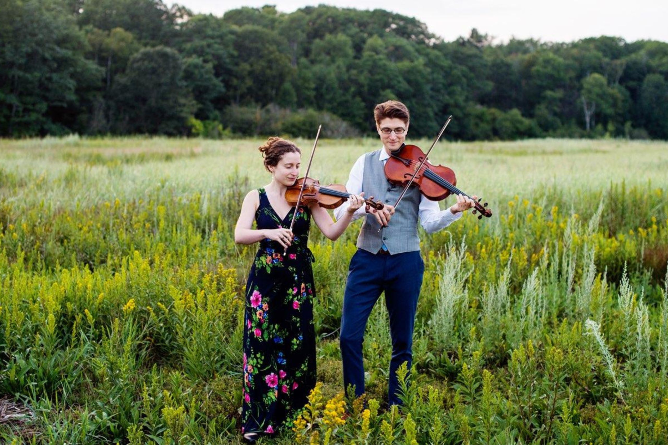 two musicians one playing a violin and another playing a viola in a field of wild flowers with hills and trees behind them.