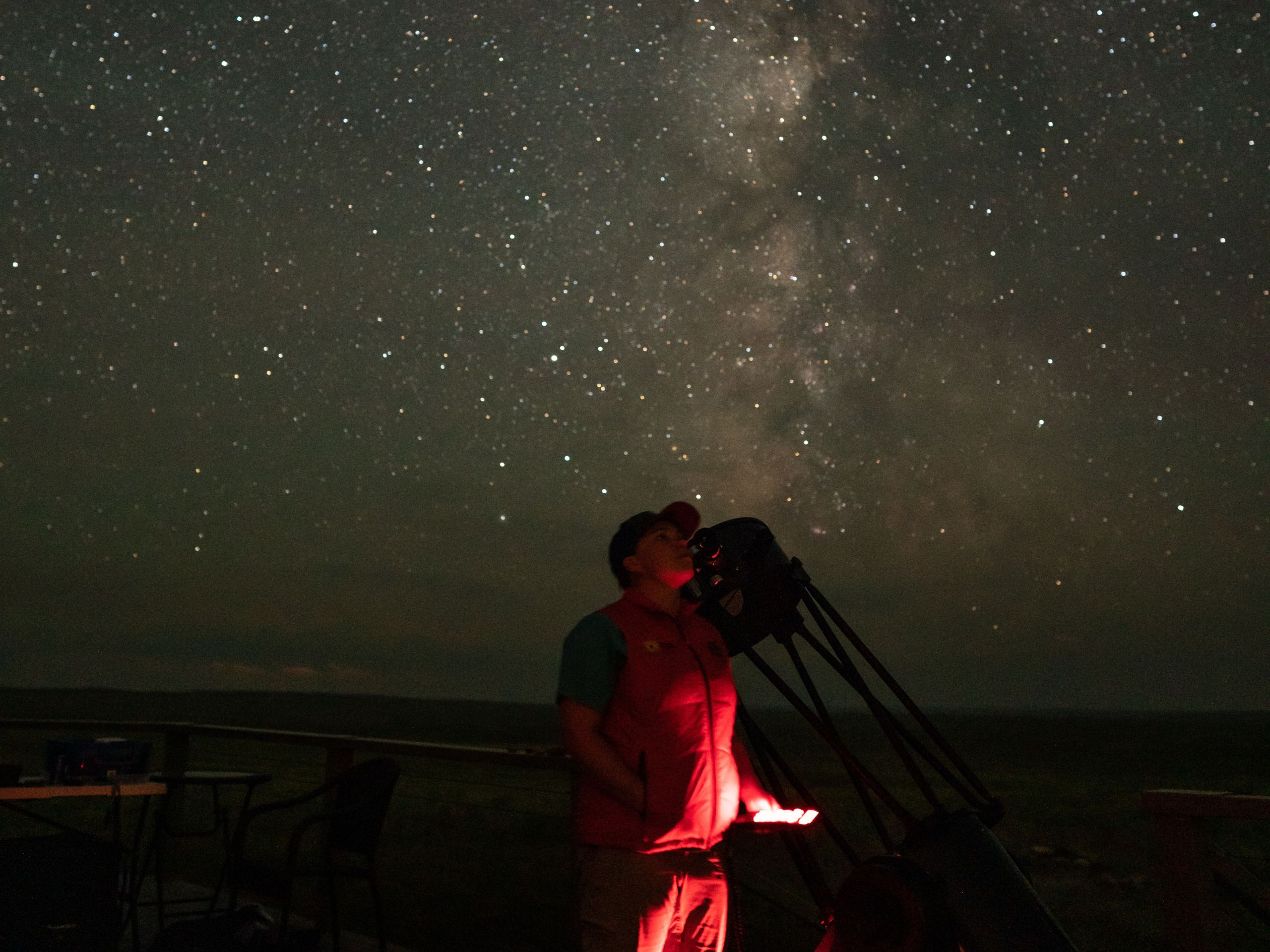 Starry sky on dark night with young person in red sweatshirt looking through a large telescope.