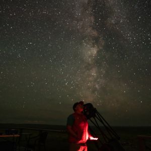 Miky way and other stars on a dark night being observed by a young person in red looking through a telescope.