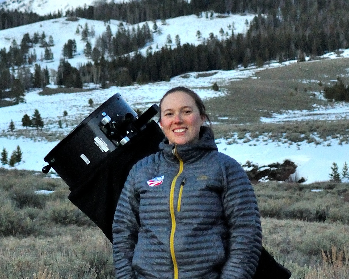 woman smiling in a puffer jacket in front of a large telescope working with fields of snow and pines behind the astronomer.