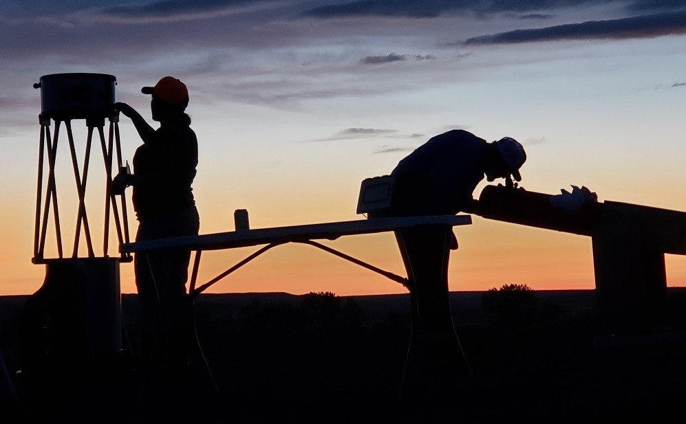 two silhouettes of people looking through telescopes at sunset.