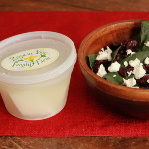 Pumpkin Vine Family Farm feta cheese in a plastic container with label with logo beside a small salad with greens, dried cranberries and feta sprinkled on top in a wooden bowl all on a red placemat.