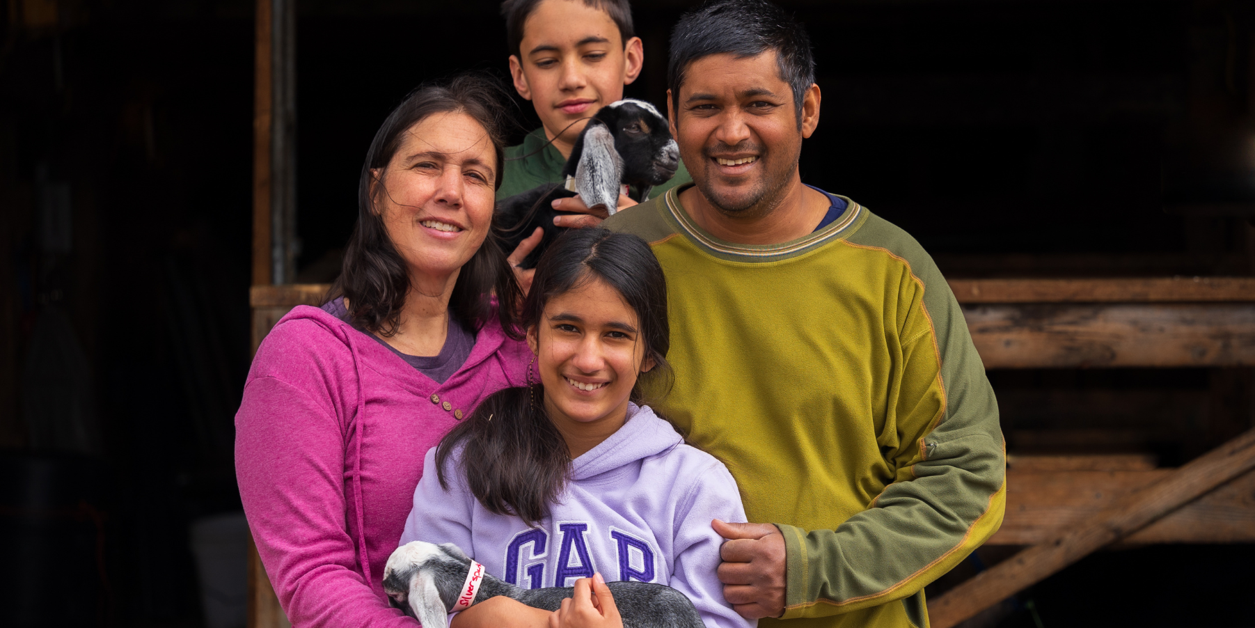 The Payson-Roopchand family owners of Pumpkin Vine Family Farm in Somerville Maine standing in front of their barn holding two baby goats.