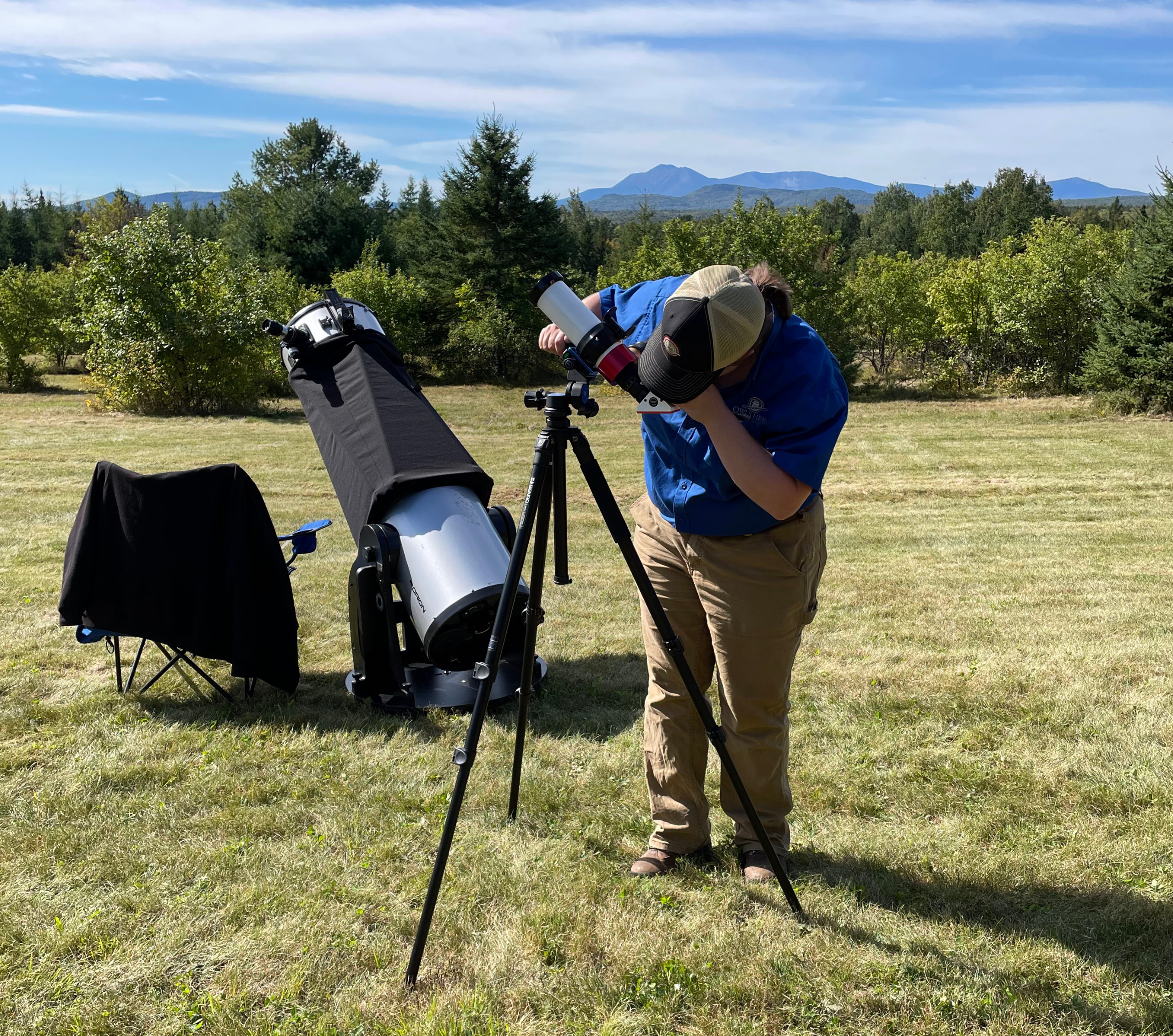 Local astronomer Rebecca Spitz, of In Focus Astronomy, in a blue shirt and brown pants with her solar telescope viewing the sun from the green fields of Pumpkin Vine Family Farm with blue skies and hills in the background.