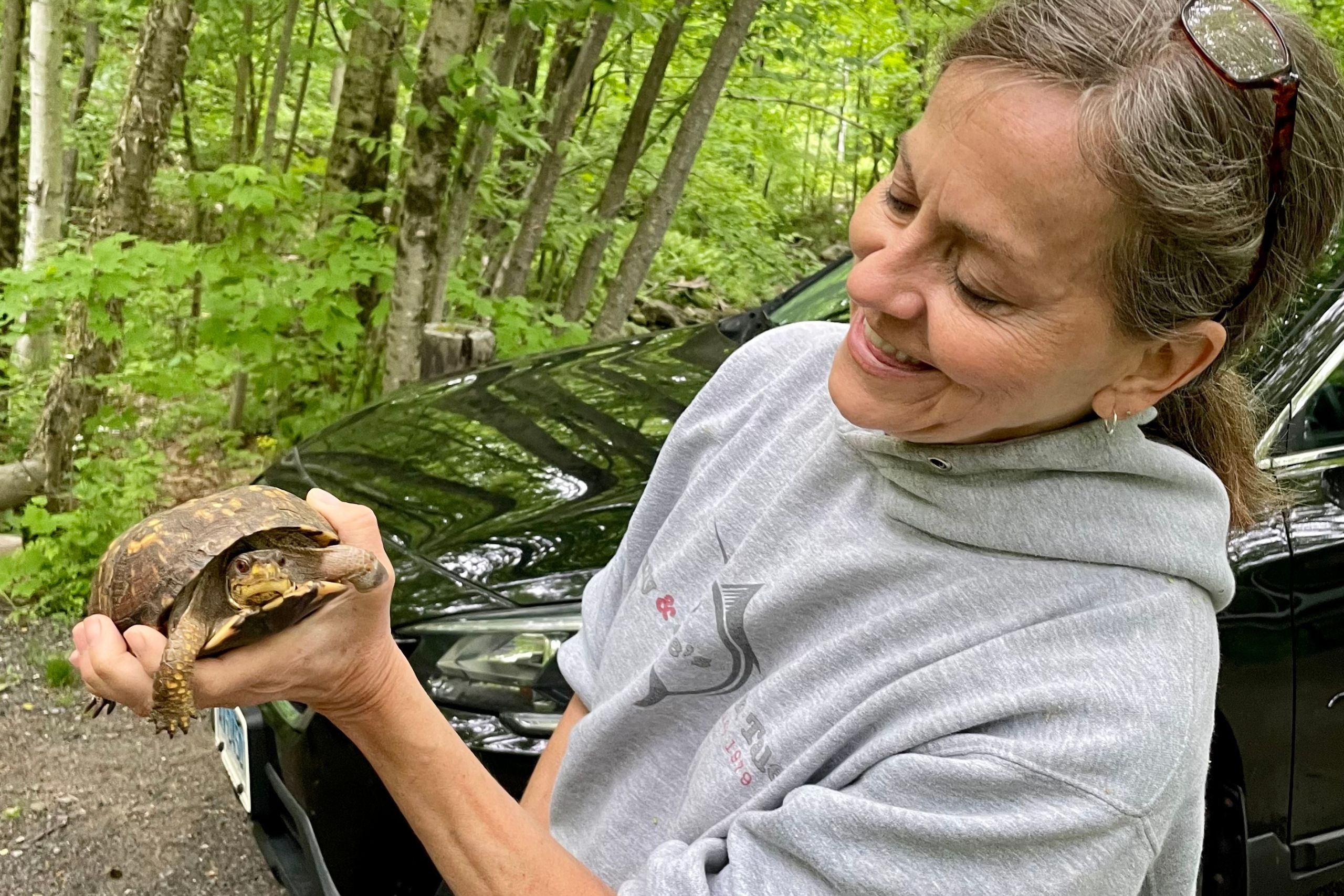 Woman in gray sweatshirt holding a small turtle and smiling at it.